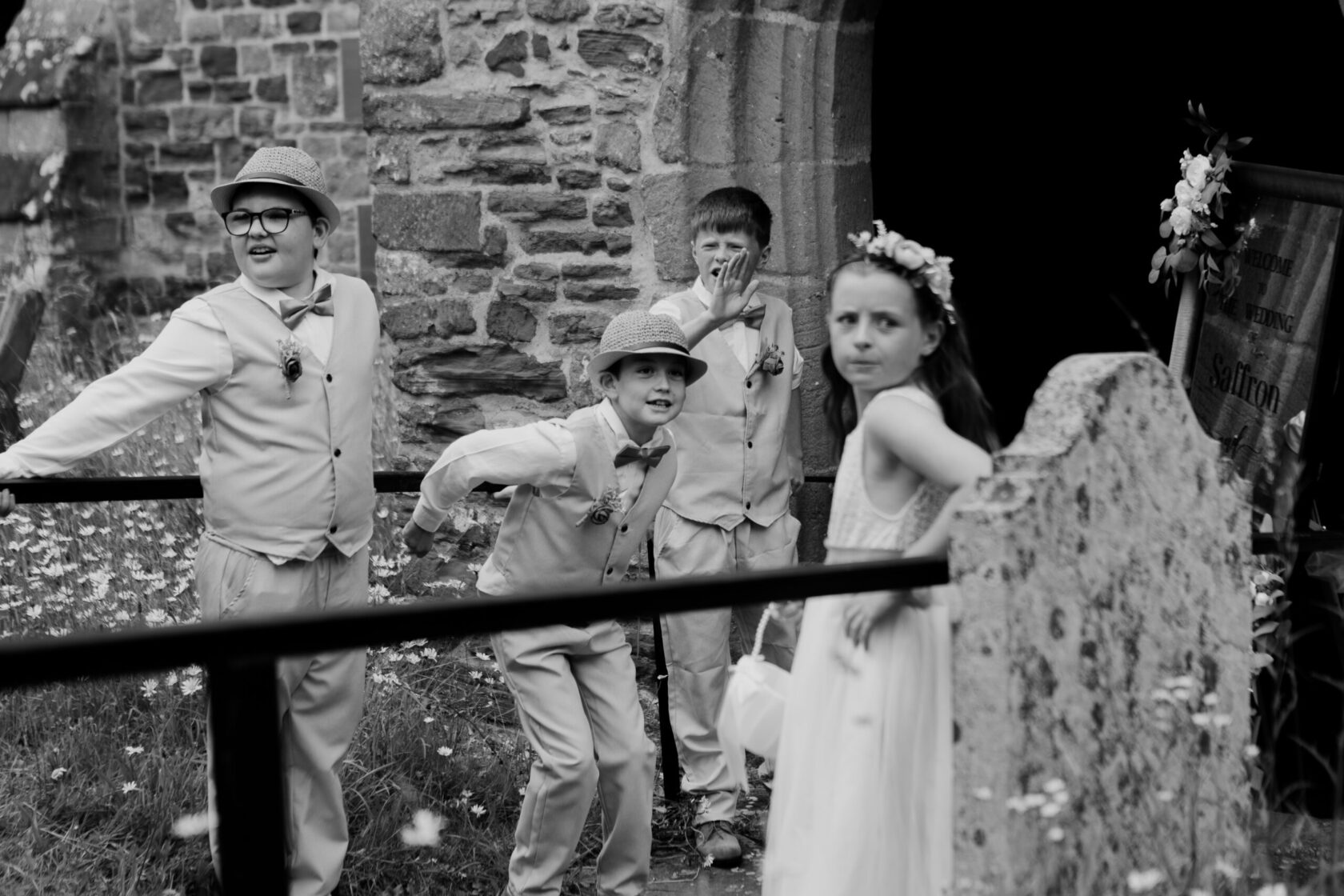 Four children dressed in formal attire for a wedding, two boys wearing hats and bow ties, one girl in a flower crown, standing near a stone wall and a gate, with daisies in the foreground.