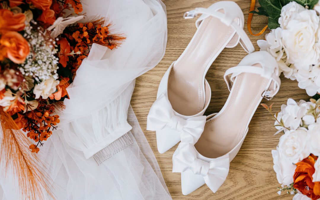 A pair of white bridal heels with bows, beside a bouquet of orange and white flowers and a veil with comb.