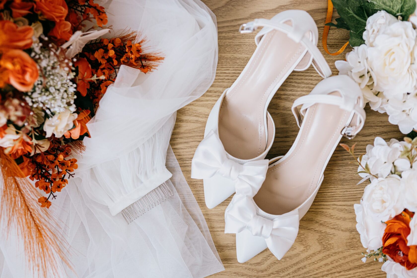 A pair of white bridal heels with bows, beside a bouquet of orange and white flowers and a veil with comb.