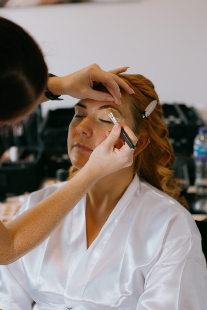 A woman with red hair wearing a white robe while a makeup artist applies eyeshadow to her closed eyes.