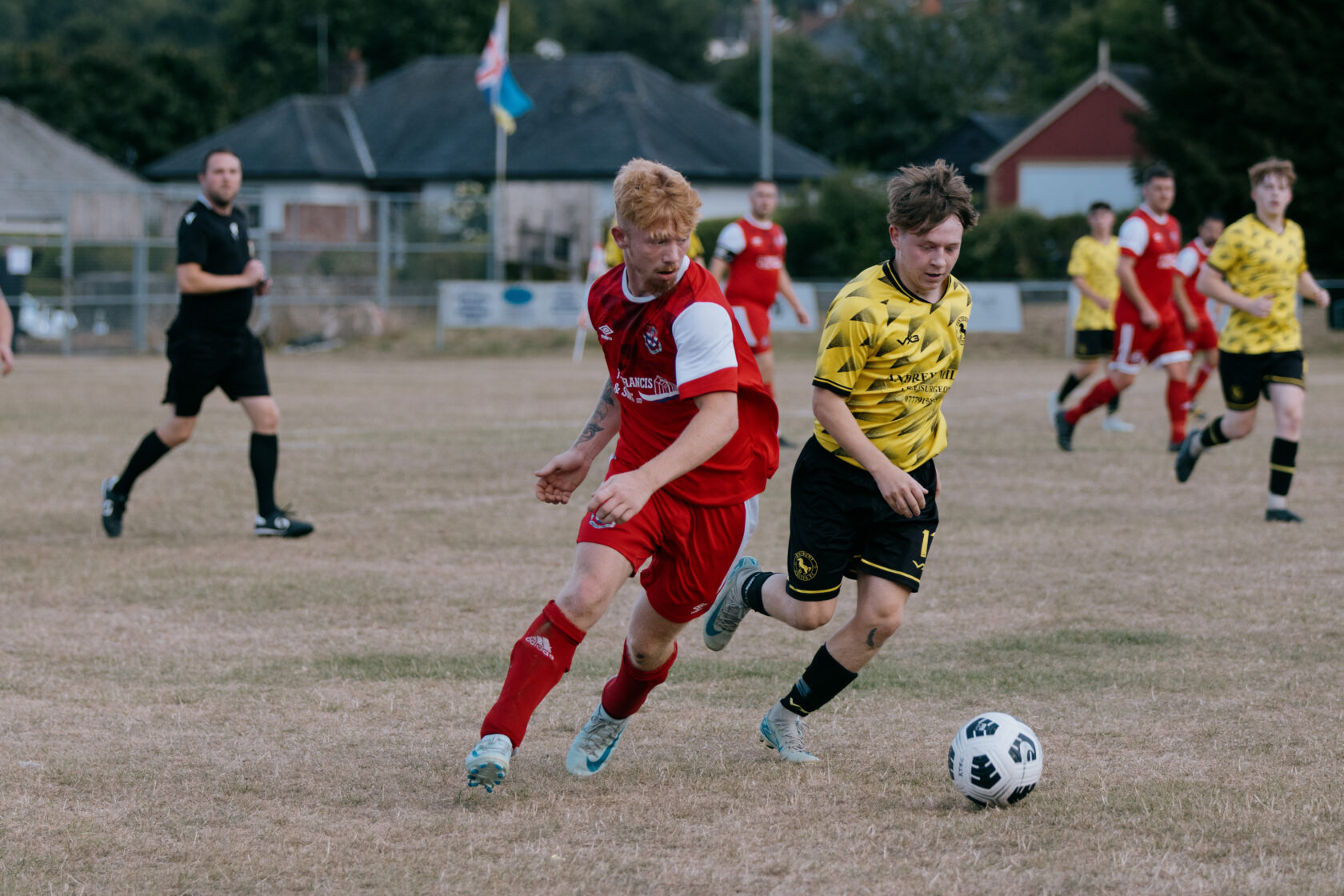Exciting Moment in Local Football Match Two football players competing for the ball on a grassy field during a match. One player in a red kit and another in a yellow and black striped kit. A referee and other players are in the background.