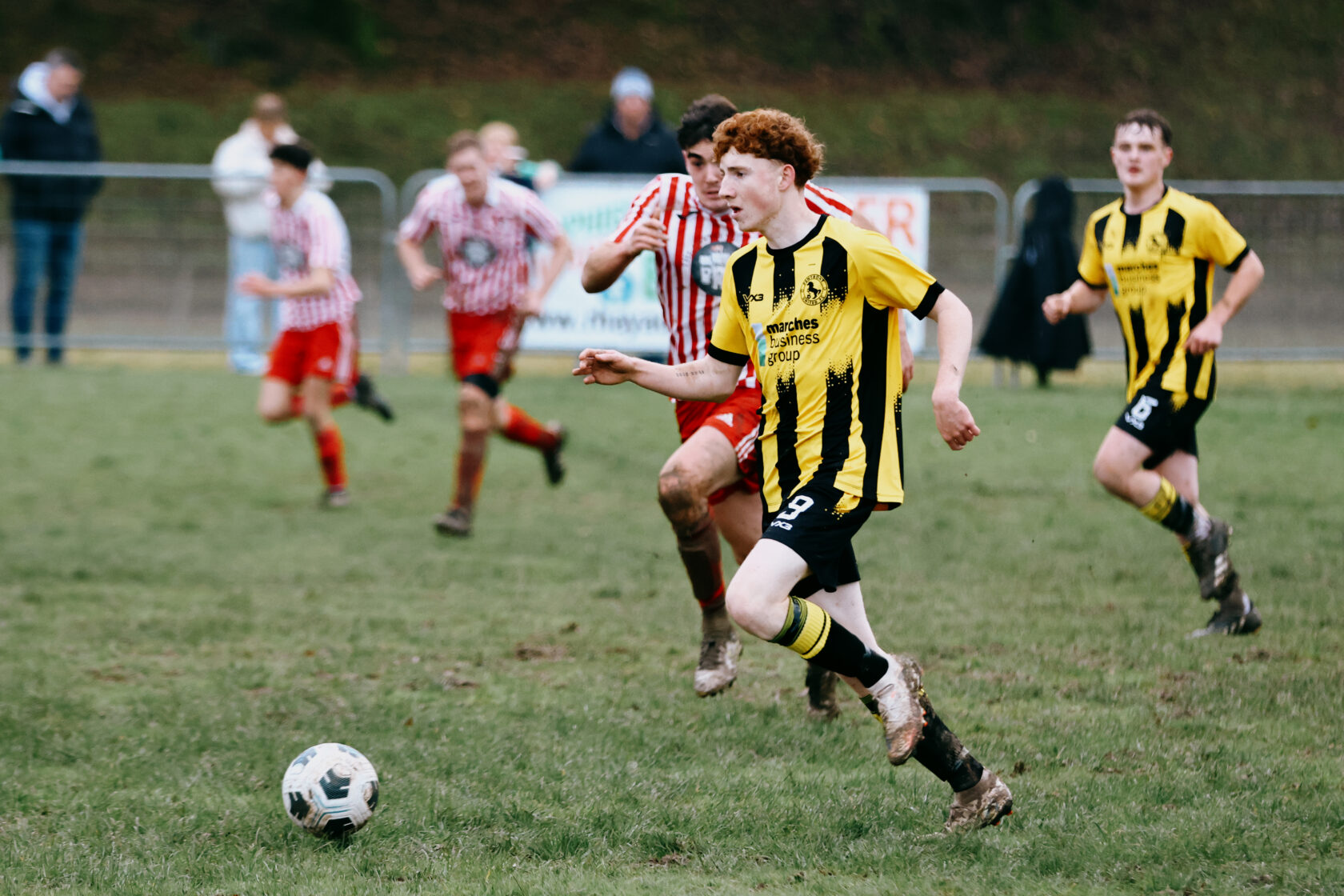 Youth Football Match Action Shot A young football player in a black and yellow striped kit runs with the ball on a muddy pitch, while players in red and white striped shirts chase behind him.
