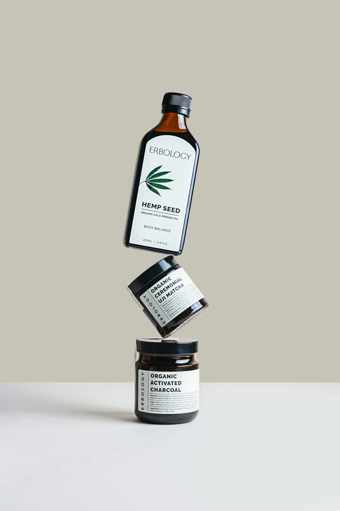 Erbology Organic Health Products Display Three Erbology health products arranged in a floating stack, featuring a bottle of organic hemp seed oil at the top, a jar of organic ceremonial uji matcha in the middle, and a jar of organic activated charcoal at the bottom. The background is a light beige colour.