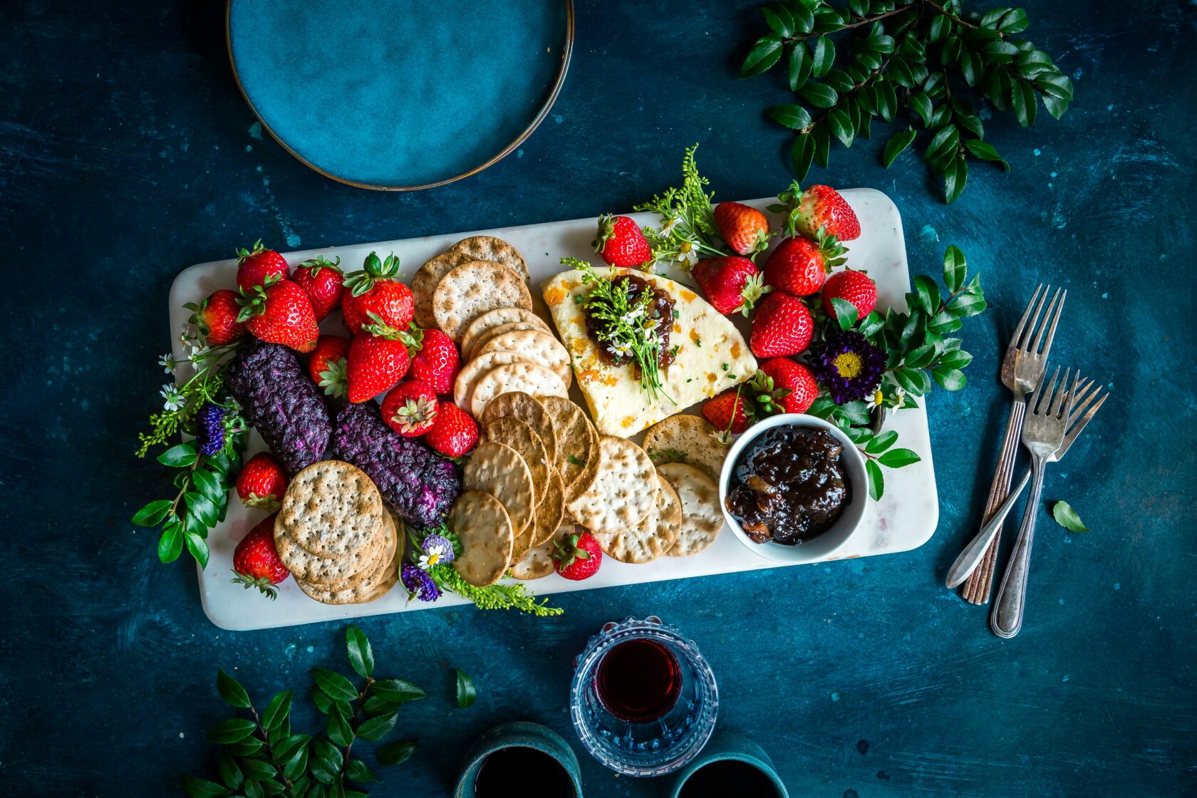 Delicious Cheese and Cracker Platter with Fresh Strawberries A beautifully arranged platter featuring slices of cheese, assorted crackers, fresh strawberries, a small bowl of dark fruit jam, and decorative herbs, all on a blue textured background.