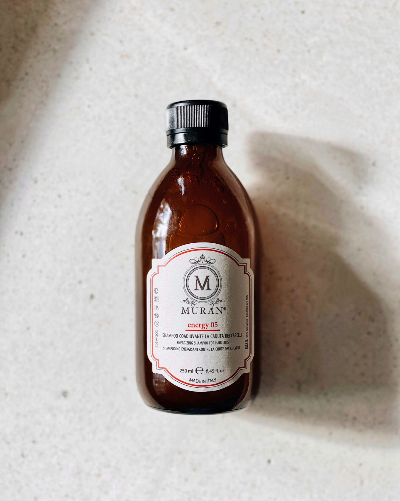 Muran Energizing Shampoo Bottle Brown glass bottle containing Muran energy 05 energizing shampoo for hair loss, labelled in English and Italian, set against a light textured surface