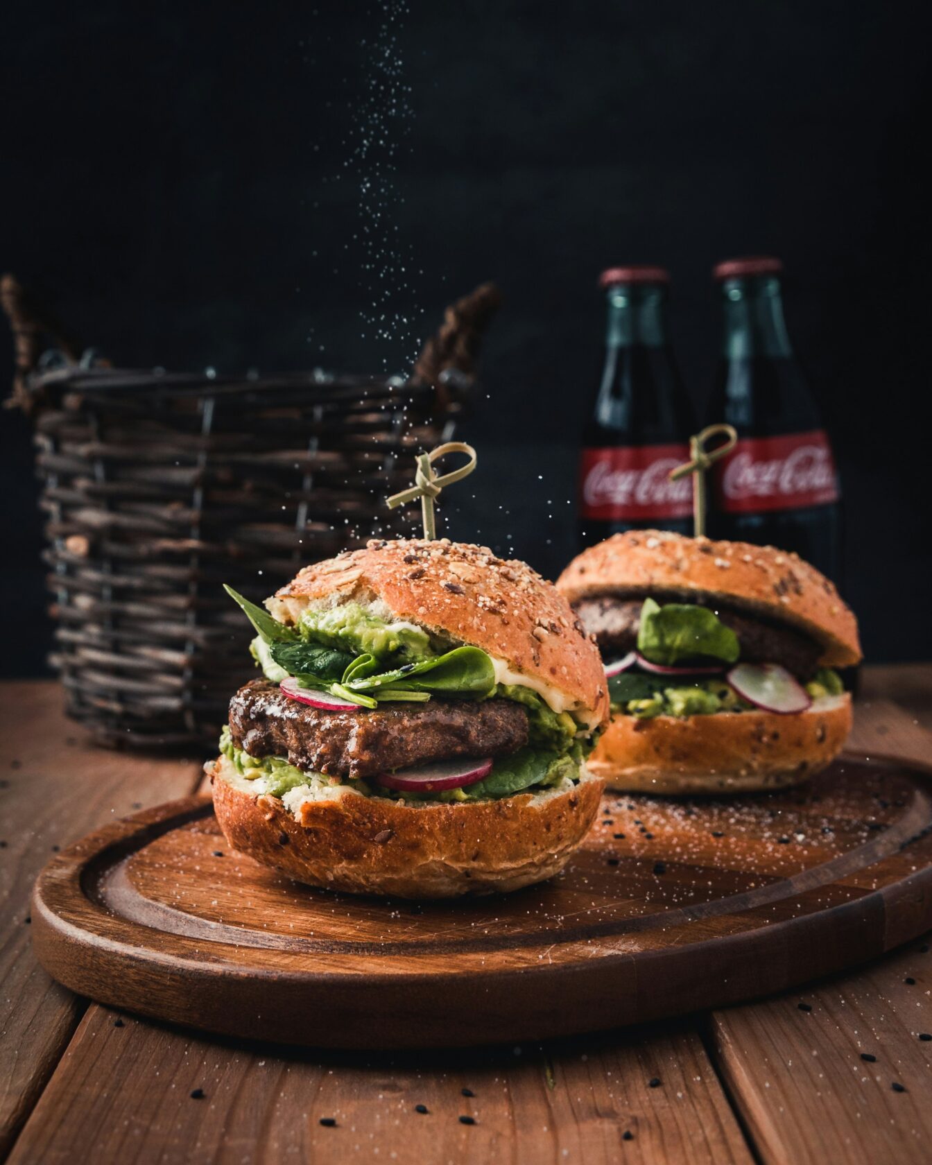 Delicious Gourmet Burgers with Coke Two gourmet burgers featuring fresh ingredients like lettuce, radish, and avocado on a wooden platter, with bottles of Coca-Cola in the background and seasoning being sprinkled.
