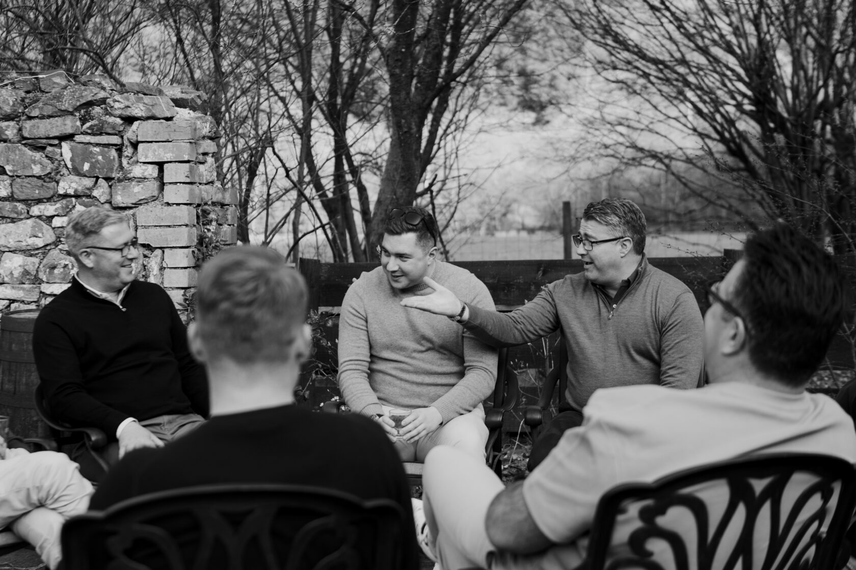 Casual Conversations Among Friends Group of men engaged in a relaxed discussion outdoors, with a stone wall and trees in the background. Two men are smiling and talking, while others listen attentively.