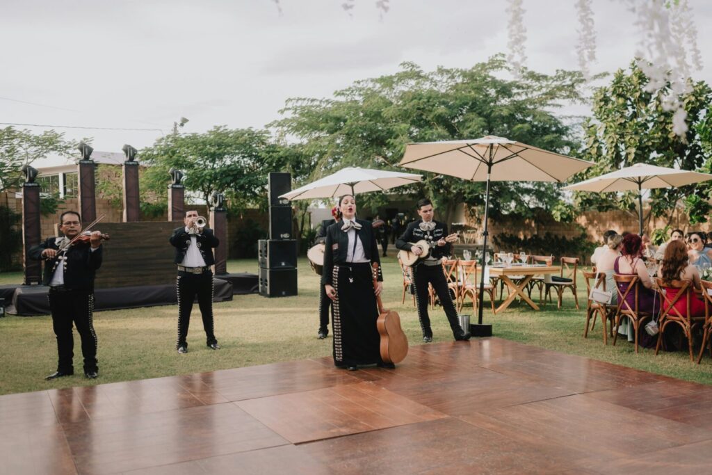 A mariachi band performing in a garden setting, featuring musicians playing a violin, trumpet, guitar, and vocals. Guests are seated at wooden tables under umbrellas.