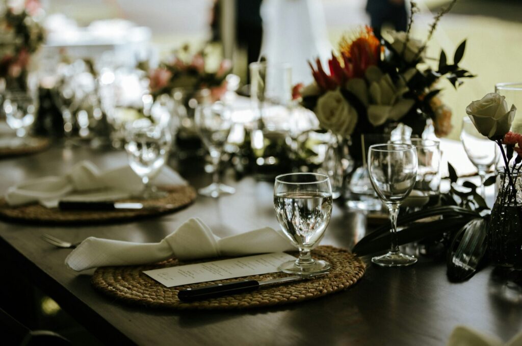 A beautifully arranged dining table with woven placemats, elegant glassware, folded napkins, cutlery, and floral centrepieces.