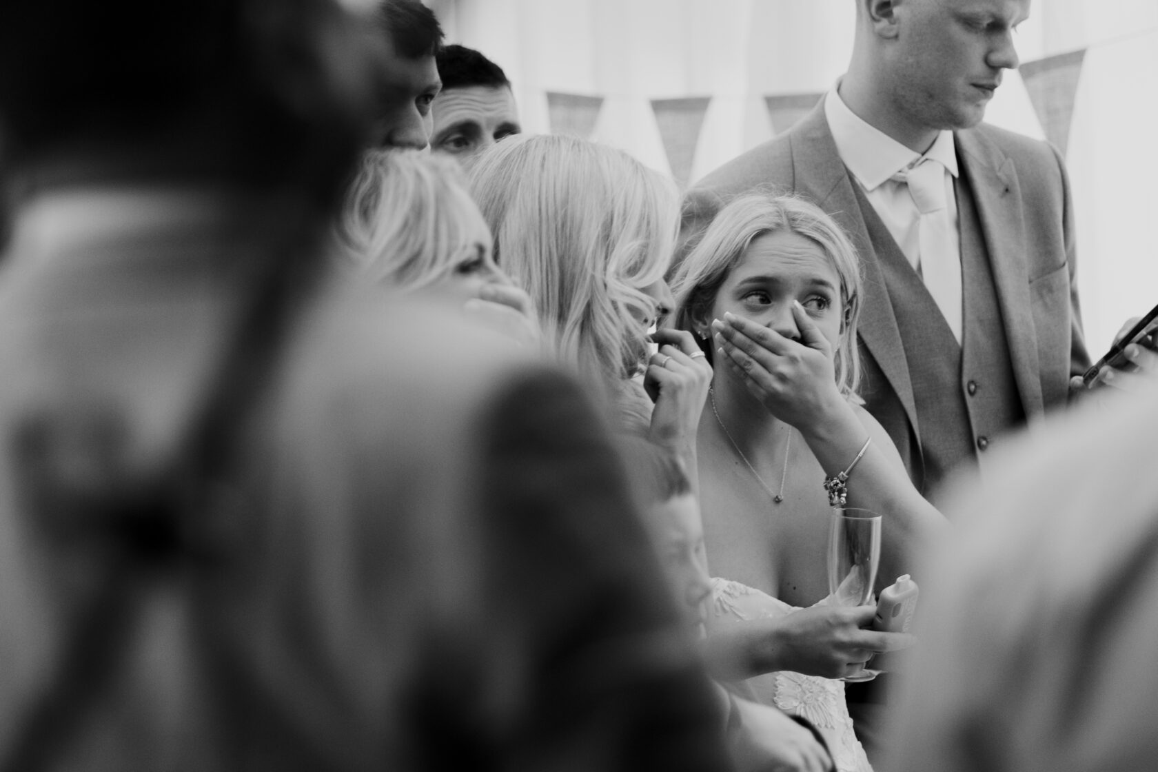 Emotional Wedding Moment A close-up of several guests at a wedding displaying emotional reactions. A bride, with her hands covering her mouth, appears to be captivated by an unexpected moment, while others around her exhibit similar expressions. Shot for Lydia Turner.