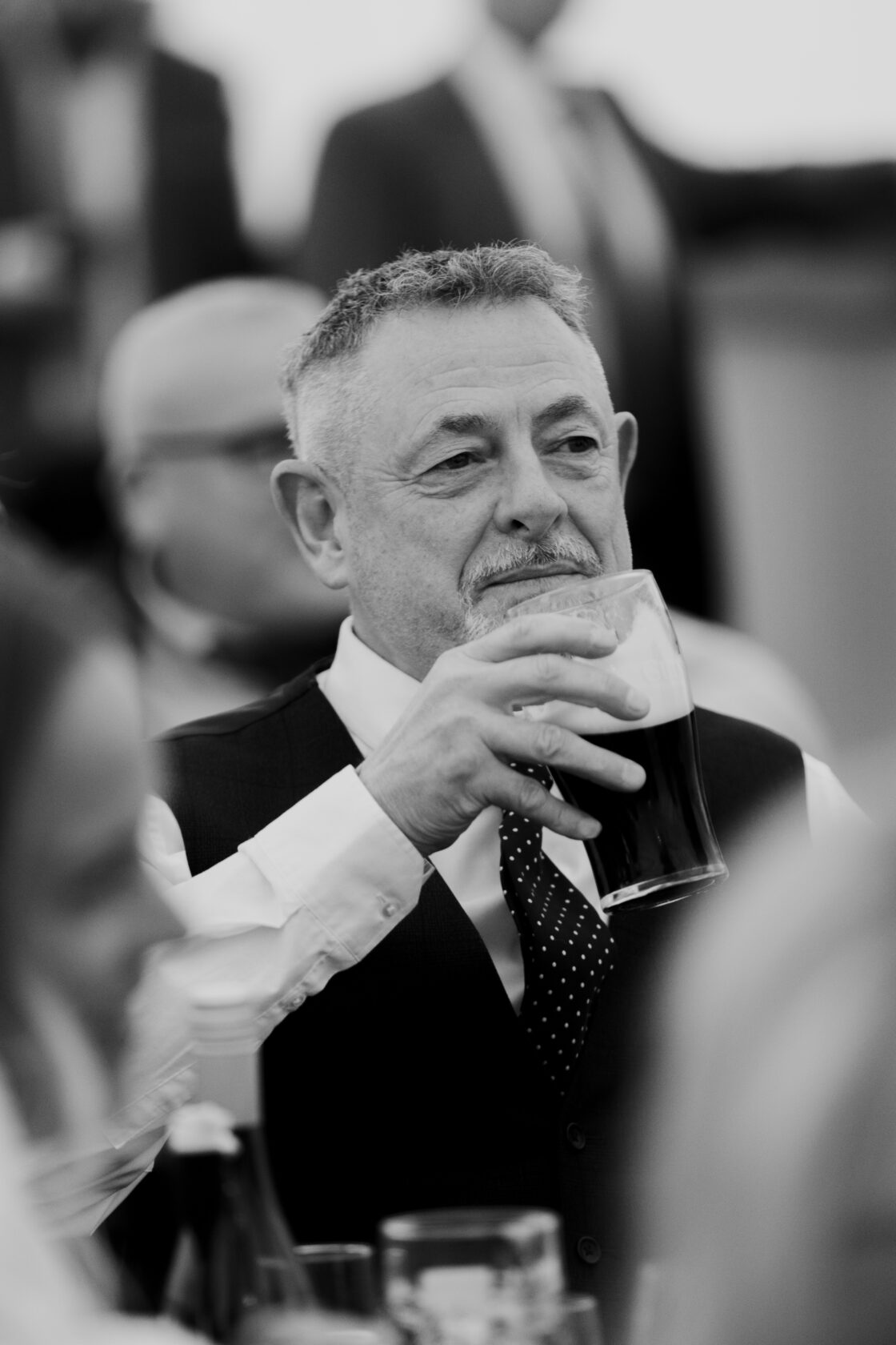 Man Enjoying a Pint in Formal Attire A middle-aged man with grey hair wearing a black vest and a white shirt with a polka dot tie, holding a pint of dark beer, smiling at an event. Shot for Lydia Turner