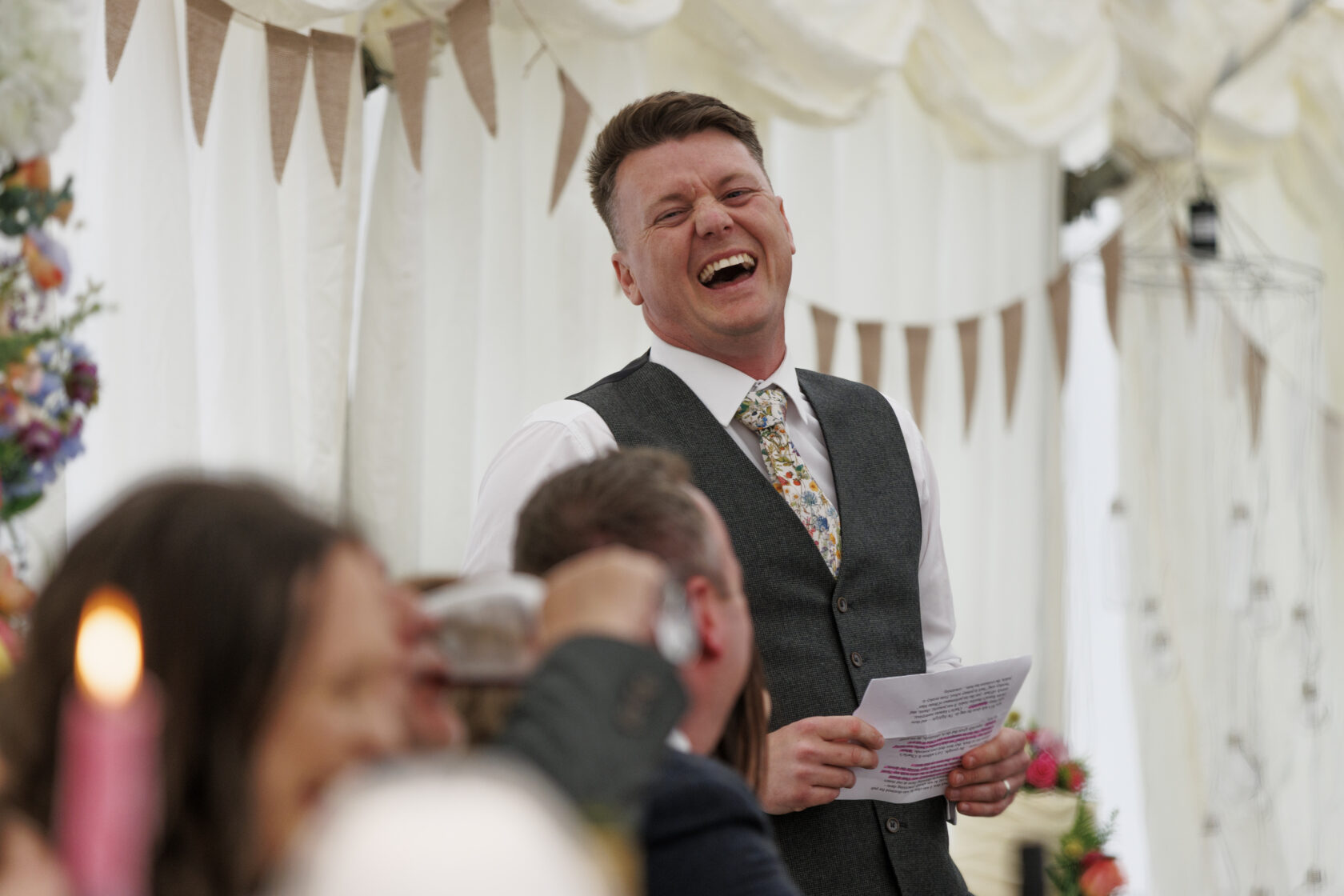 Joyful Wedding Speech in a Marquee A smiling man in a grey waistcoat and floral tie delivers a speech at a wedding, with guests laughing and enjoying the moment. Shot for Lydia Turner.