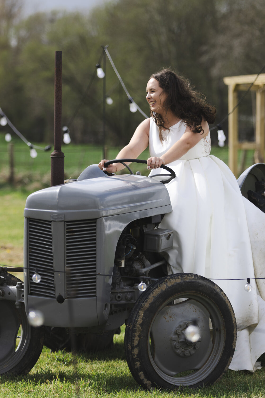 Bride Joyfully Riding a Vintage Tractor A joyful bride in a white wedding dress, laughing while driving a vintage grey tractor in a scenic outdoor setting with fairy lights in the background. Shot for Lydia Turner