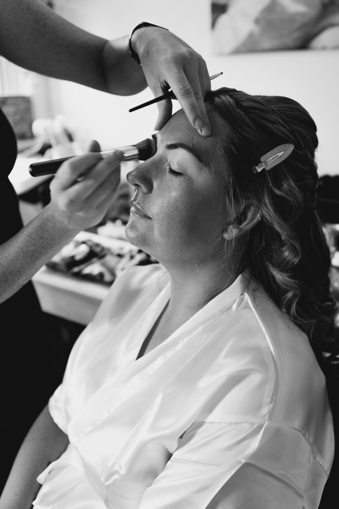 Bridal Makeup Preparation A woman sitting with her eyes closed while a makeup artist applies foundation to her face, with her hair in soft curls and wearing a white satin robe.