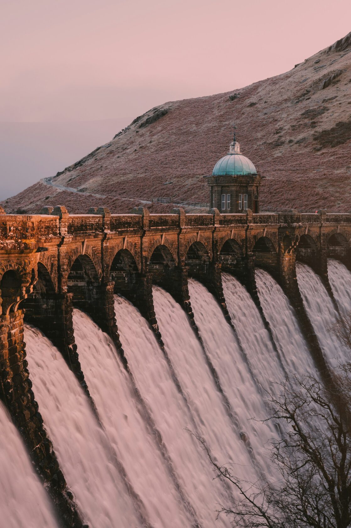 A dramatic view of a historic stone dam with multiple water outlets flowing into a river below. A small domed tower sits atop the dam, surrounded by a hilly landscape under a pastel sky.