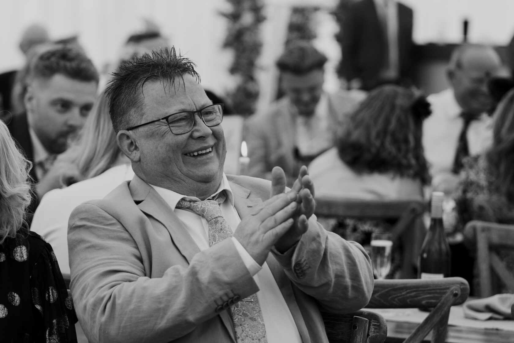 Joyful Celebration at a Wedding Party A man with glasses in a light grey suit and patterned tie claps and smiles, surrounded by fellow guests at a wedding reception. Shot for Lydia Turner.