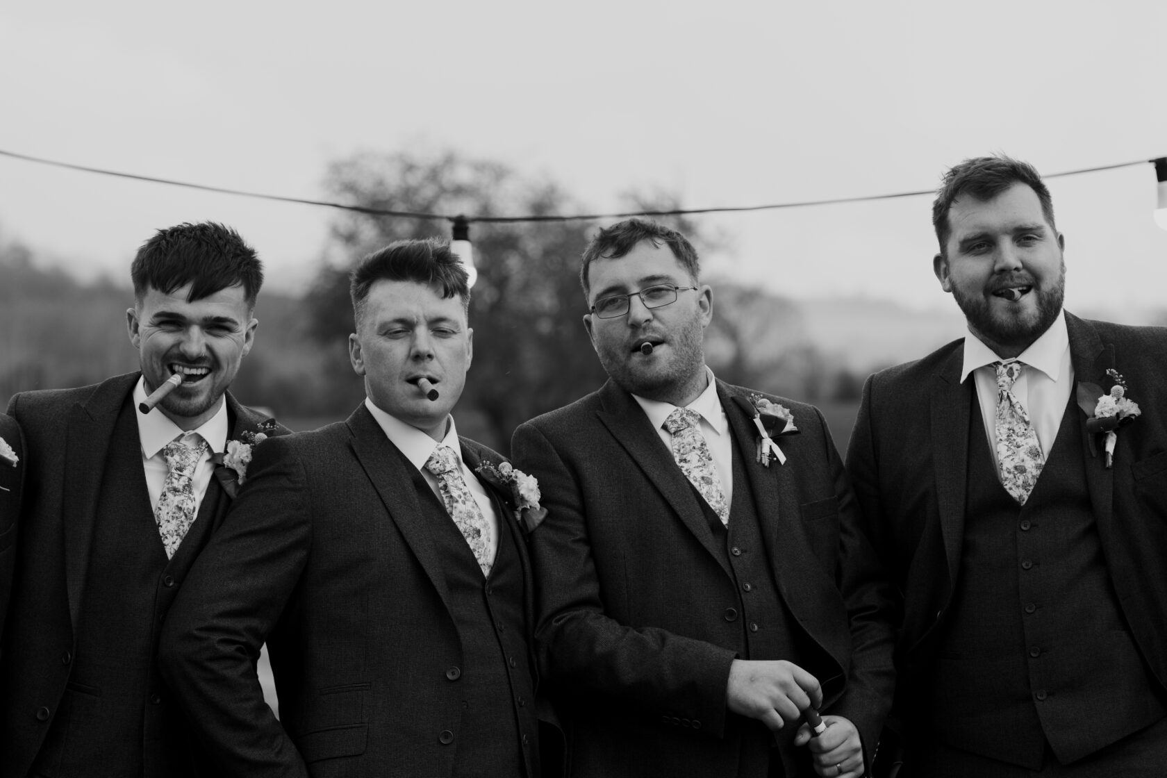 Groom's Party in Stylish Suits and Cigars Four men in formal suits laughing and posing together, each holding a cigar, with floral ties and boutonnieres. Shot for Lydia Turner.