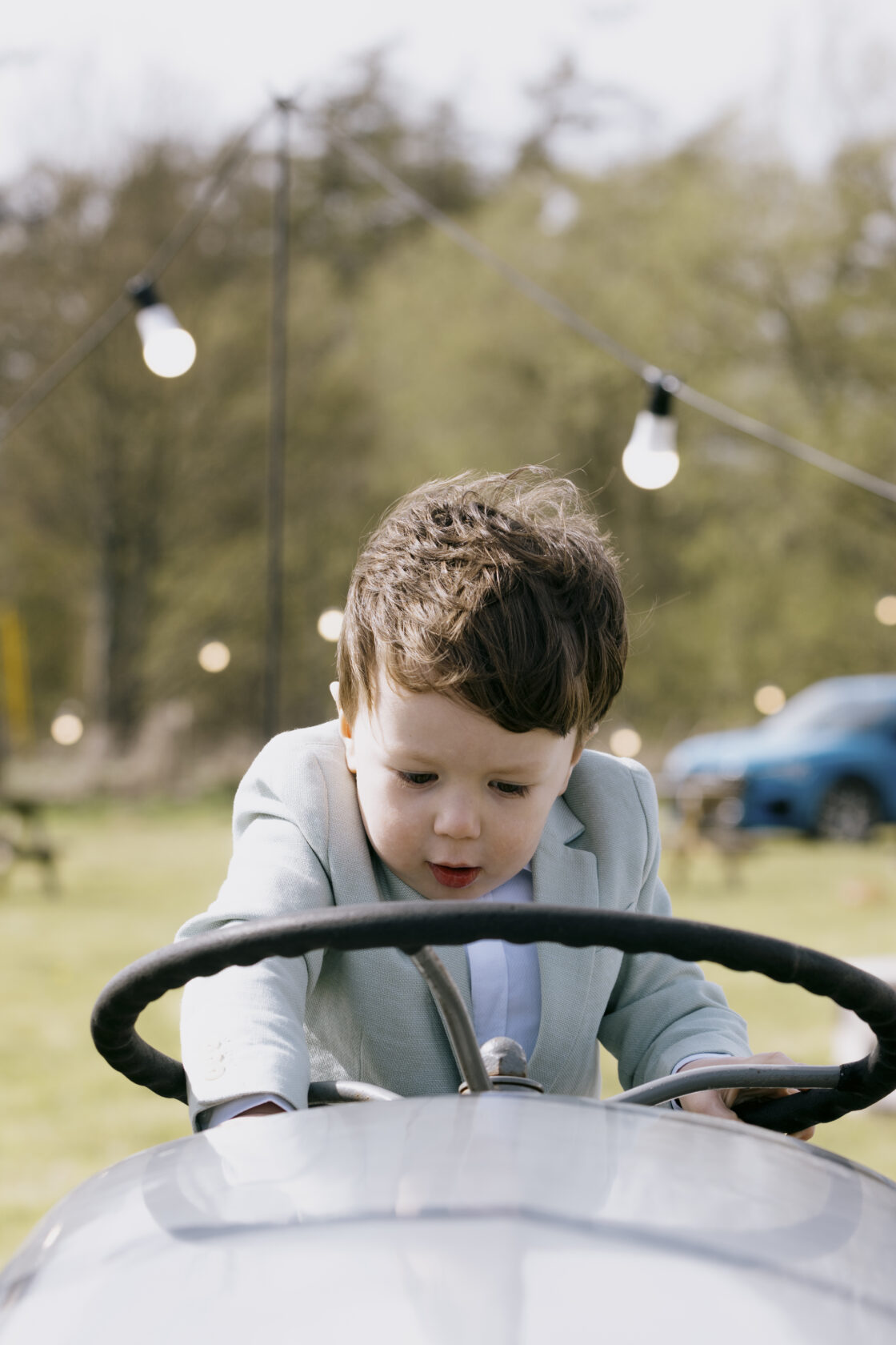 Young Boy Driving Classic Toy Car A young boy in a light blue suit leaning over the steering wheel of a classic toy car, focused and engaged. Soft background with greenery and hanging lights. Shot for Lydia Turner.