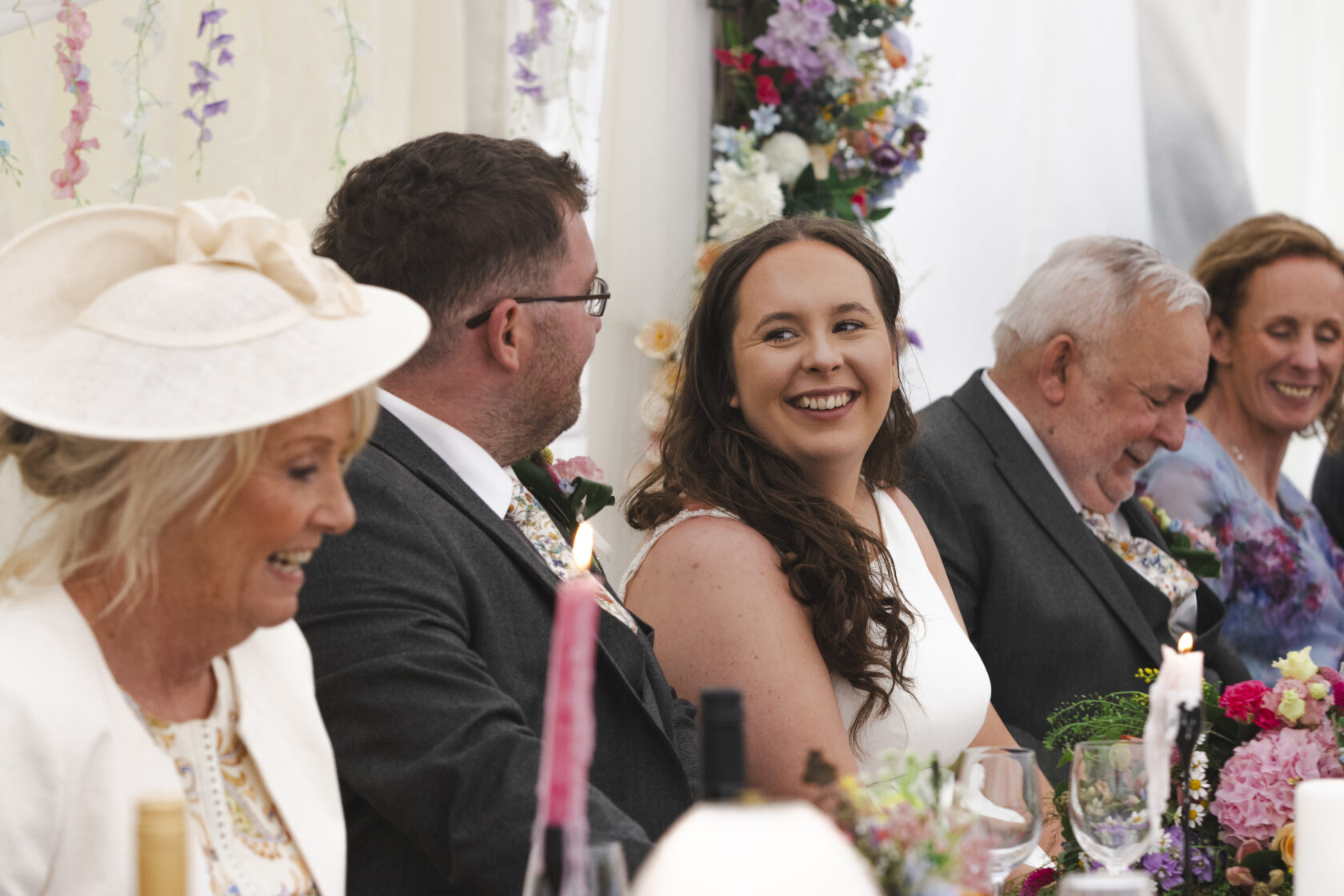 A woman in a white dress smiles at a seated man at a wedding reception, surrounded by friends and family. Decorated tables with flowers and candles create a festive atmosphere. Shot for Lydia Turner.