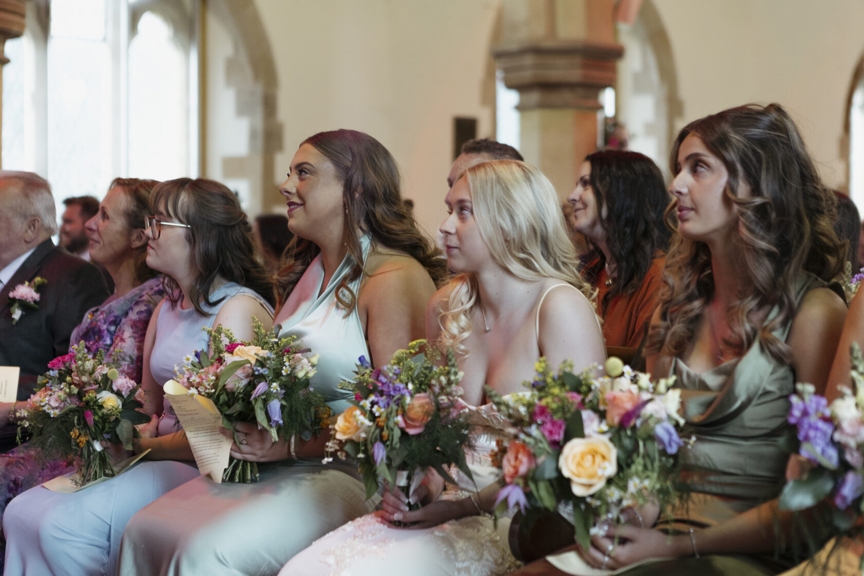 A group of bridesmaids sitting during a wedding ceremony, holding bouquets of flowers and wearing elegant dresses in various pastel colours. Shot for Lydia Turner.