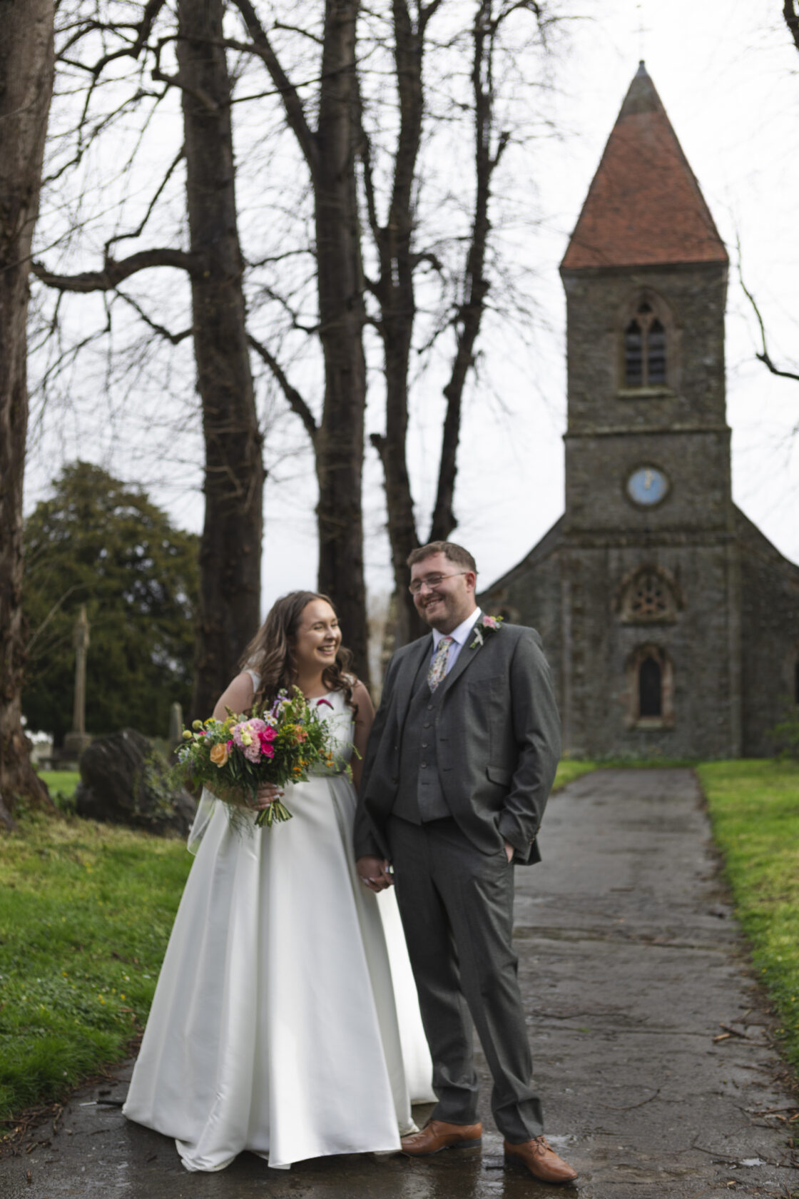 Outdoor Wedding at Historic Church A smiling bride in a white dress holds a vibrant bouquet, standing beside a groom in a grey suit outside a historic church with a clock tower, surrounded by bare trees.
