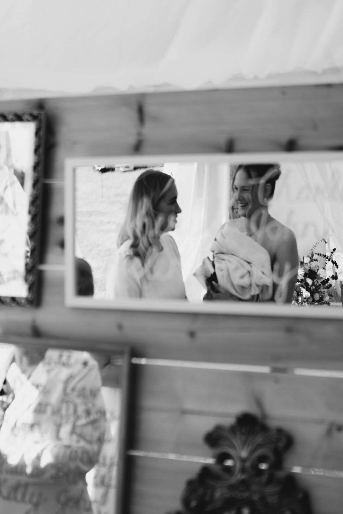 Reflection of Joy: Wedding Moments Two women, one in a strapless dress and the other in a white blouse, share a joyful moment reflected in a mirror. Flowers and decorations are visible in the background. Shot for Lydia Turner.