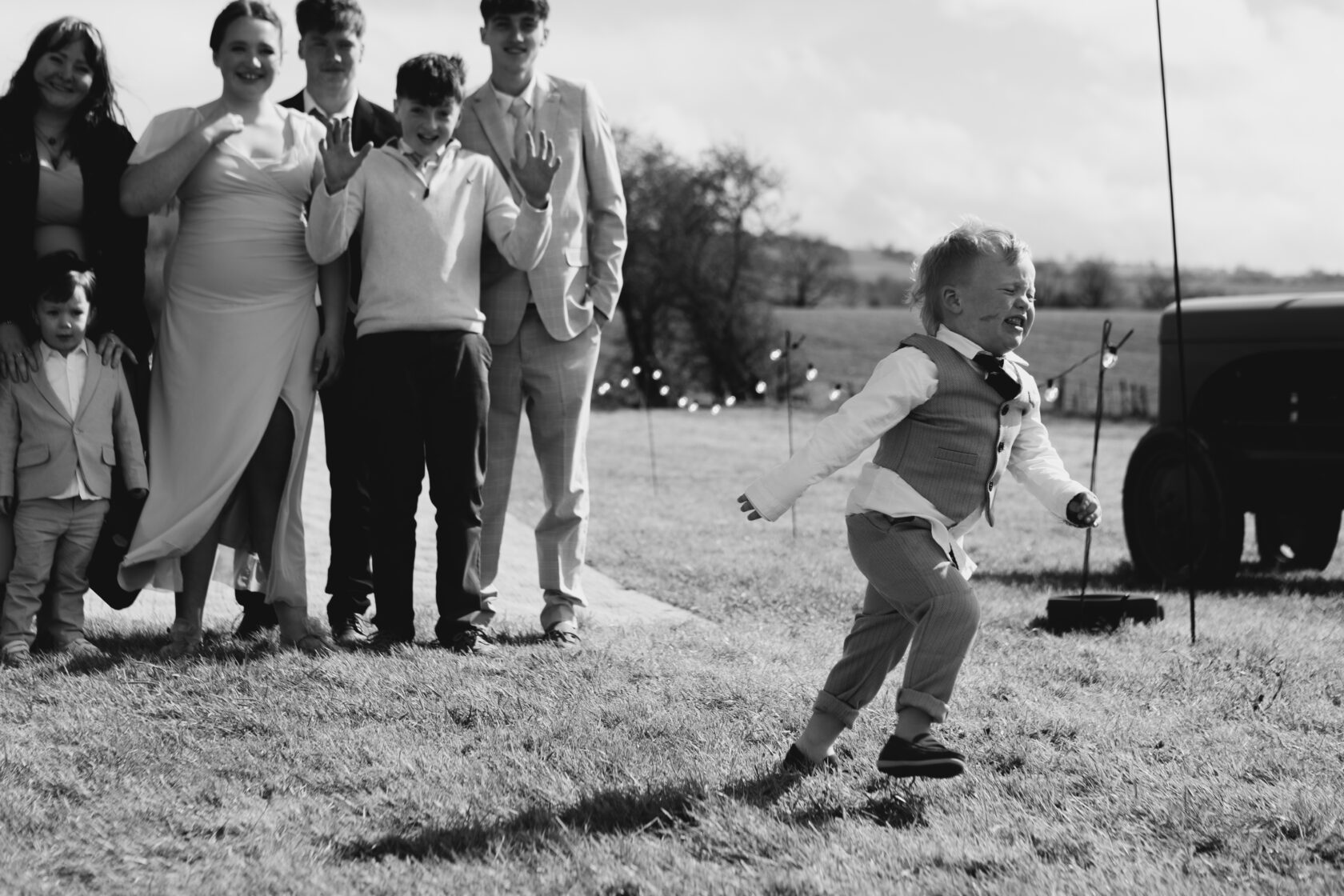 A young boy in a grey suit is joyfully running across a grassy field, while a group of smiling family members stands in the background. Shot for Lydia Turner.