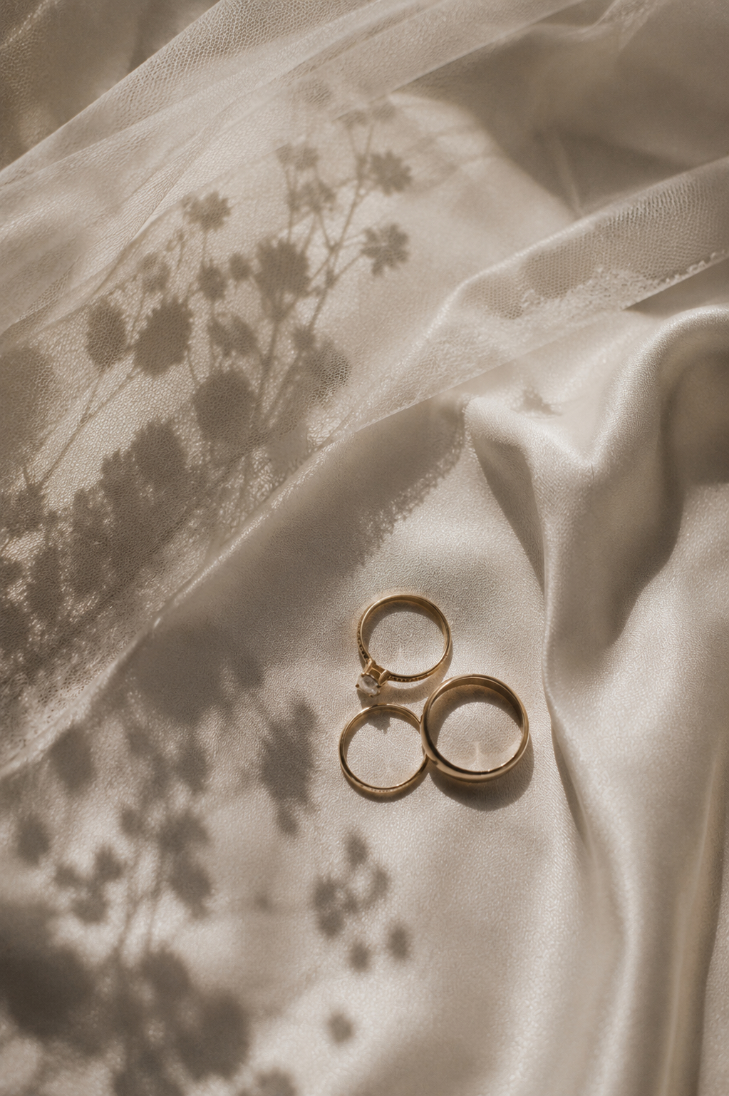 Elegant Wedding Rings on Satin Background Three gold wedding rings laid on soft satin fabric, with delicate floral shadows cast upon them. Shot for Lydia Turner